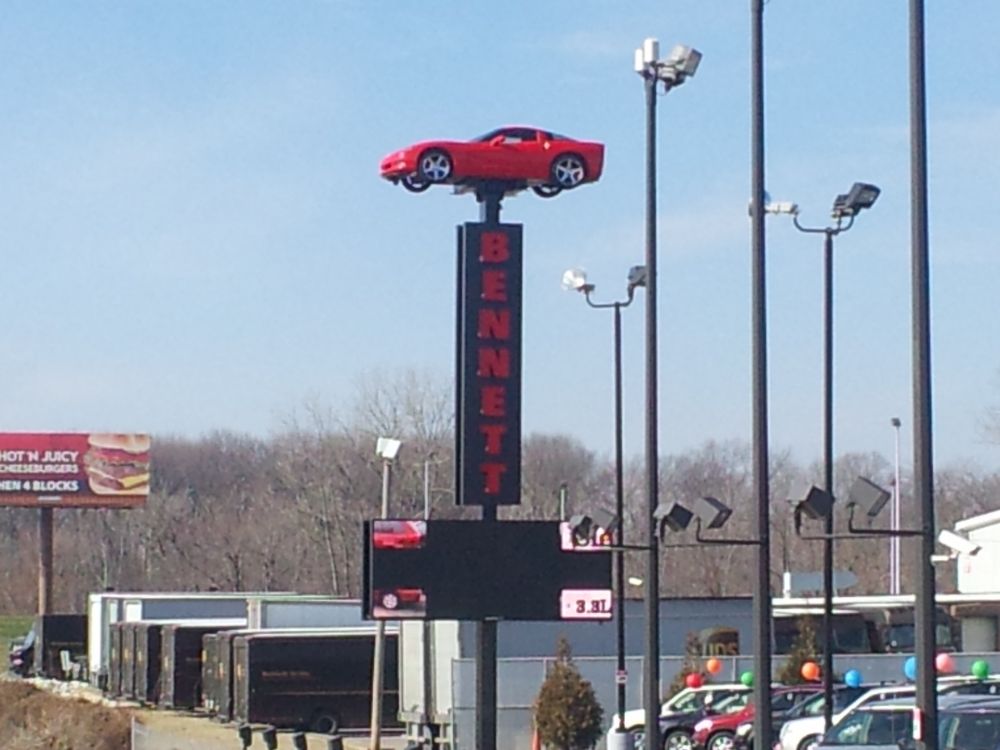 Sign at a car lot, topped by a red Corvette. Below the car, the name "Bennett" is spelled-out, vertically. Beneath that, a light sign is in the process of changing advertisements.