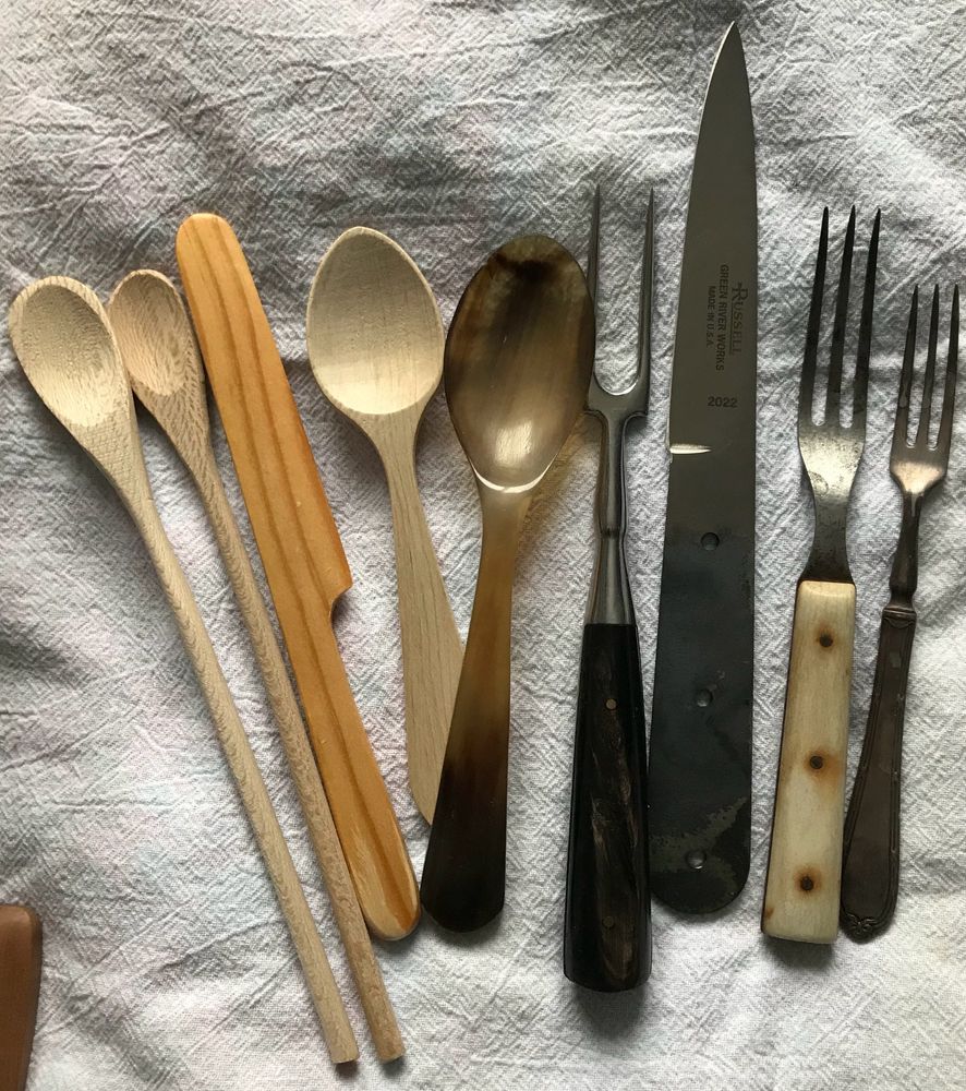 A photo of two spoons made of wood, a wooden butter knife, another wooden spoon, a spoon made of cow horn, a two-prong steel fork, an unfinished eating blade, a three-prong steel fork with a bone handle, and a small silver three-prong fork, all laid out side by side on a white cotton textured fabric.