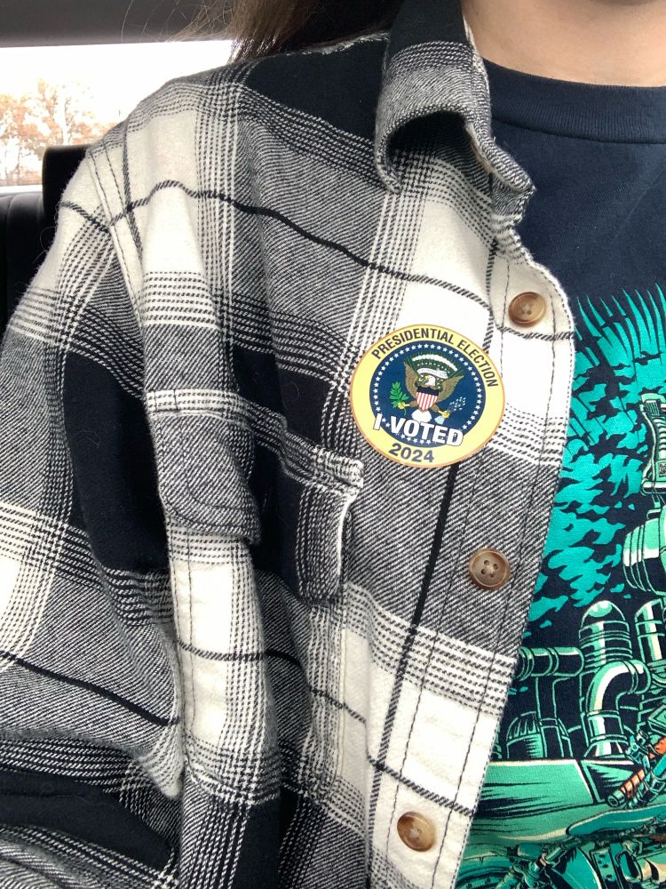 selfie of a sticker on a black and white checkered flannel shirt. the sticker reads “presidential election - I voted - 2024”