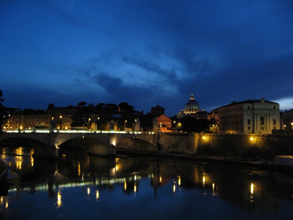 A dusk view of the Ponte Vittorio Emanuele II from the Ponte Sant’Angelo, looking toward the lit dome of St. Peter’s Basilica at the Vatican. The placid waters of the Tiber River reflect the city lights and the cobalt sky.