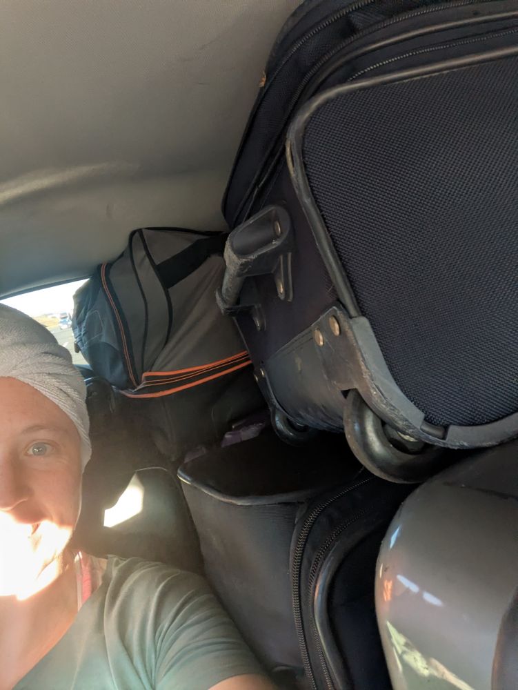 Half of Kit's face and upper body are visible. They are wearing a green shirt and a hair wrap while smiling too enthusiastically in front of their luggage Tetris'd into a car.