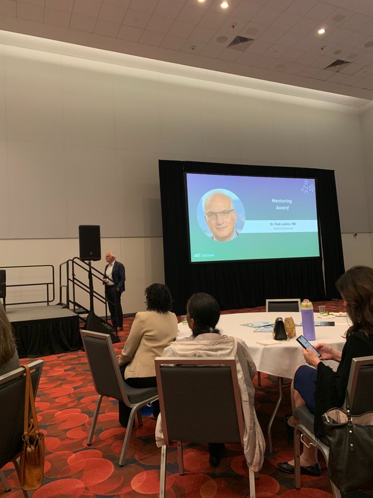 Dr. Fadi Lakkis waiting at the bottom of the stairs to enter the stage, next to a projector screen with his photo and the words "Mentoring Award"