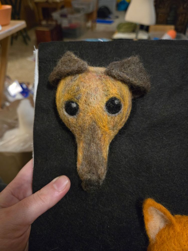 A close up view of the brindle greyhound. Her long nose ispointed downwards, with a darker brown towards the nose end. Her brown ears are flopped over her head and she has slightly anxious eyes. 