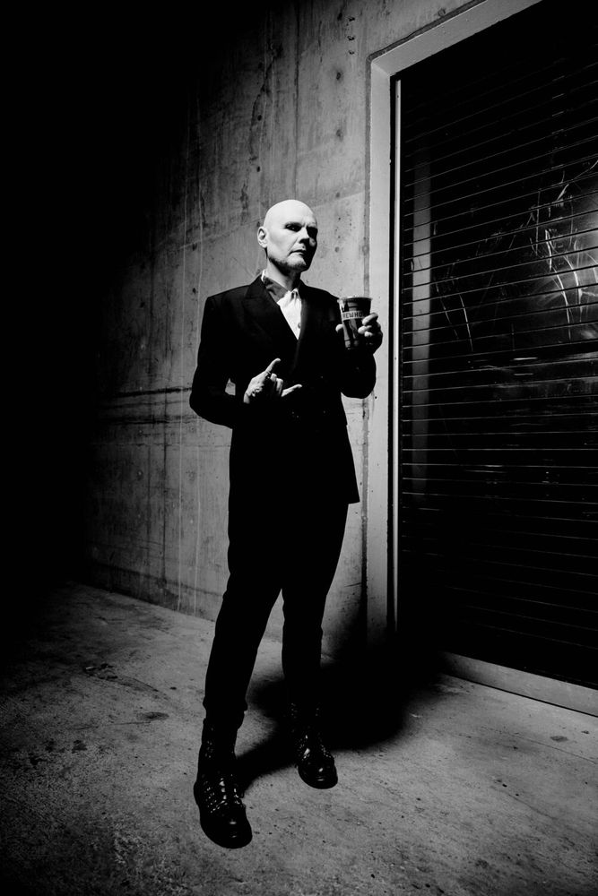 A darkly lit, suit-wearing Billy Corgan is photographed in an alley making a metal fist in one hand and holding a dark Erewhon smoothie in the other