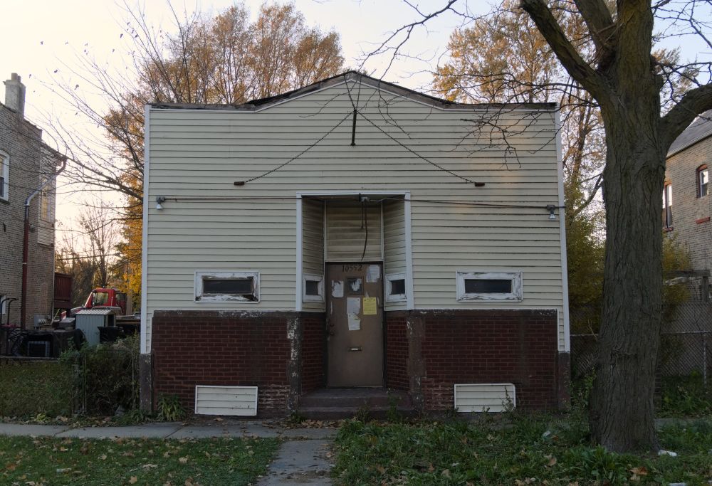 A one story storefront building, with narrow windows that suggest perhaps an old tavern, is seen with no immediate neighbor in empty lots on either side. It has a metal pole sticking out from the front of its facade where a sign used to be, and its front door is littered with advisory postings of some sort from a government agency. In the background, the edges of two two story brick buildings can be seen, and next to one of those buildings is a yard full of vehicles and various possessions.