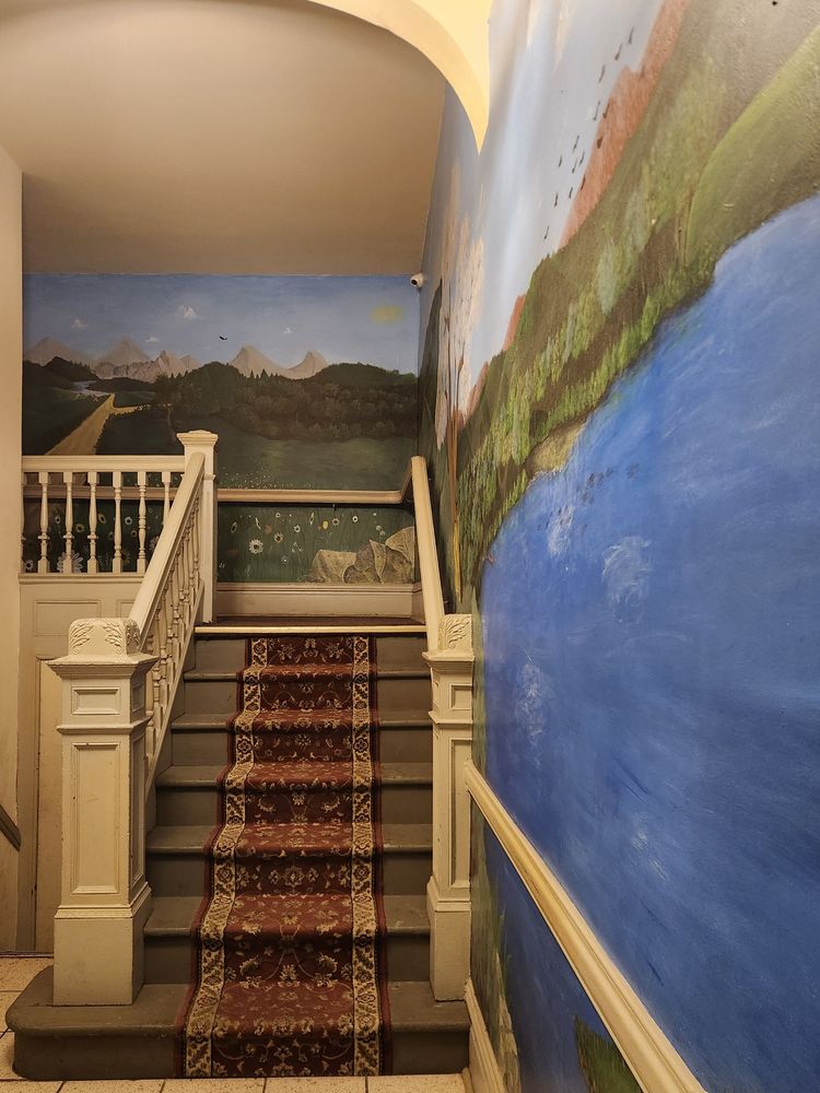 A photo of the interior of an early 20th century apartment building shows a wooden staircase leading up to a shared hallway. The staircase has been painted over and some carpeting added. Along two walls, somebody has painted a scene depicting a wide blue lake hemmed by hills and a meadow of flowers, giving way to mountains in the distance. The work appears likely to be that of a skilled hobbyist, rather than a professional muralist.
