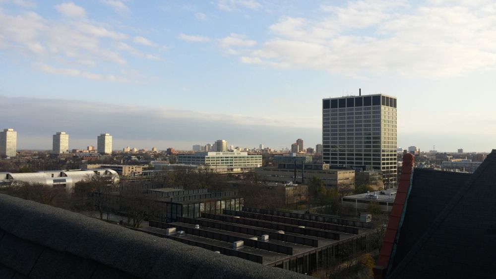 A photo taken in early evening shows sunset tones over the peaks of a roof about five stories off the ground in the foreground, and a college campus comprised mostly of midcentury buildings spread out below it and a wider cityscape leading away towards a lake further away.