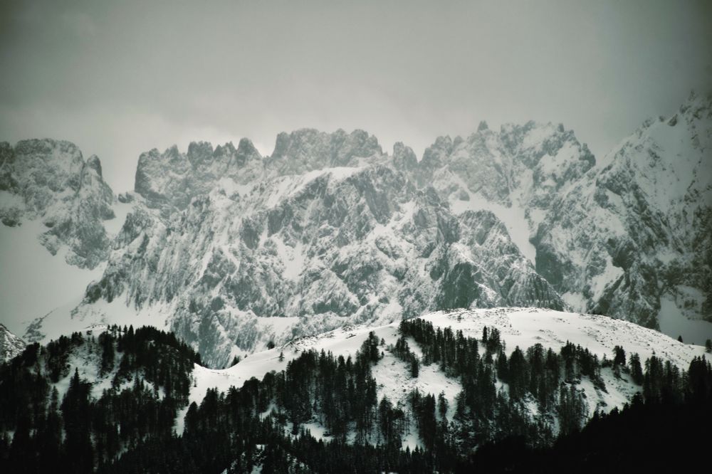 Wilder Kaiser north face in winter with Kleines Törl mountain pass visible left, snow-covered limestone peaks and forested slopes, dramatic cloudy sky, Tyrol Austria May 2019