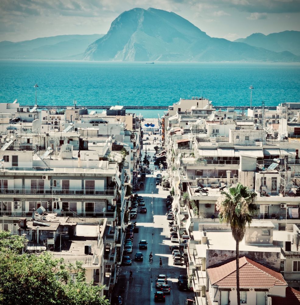 Aerial view of Patras Greece city street leading to turquoise Gulf of Patras with mountain silhouette across water, white buildings and urban architecture in foreground, June 2018