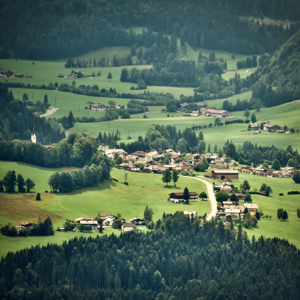 Aerial view of Schwendt village in Kaiserwinkl valley Tyrol, traditional Austrian houses scattered across green meadows, forested hills, white church visible, typical Alpine rural landscape.