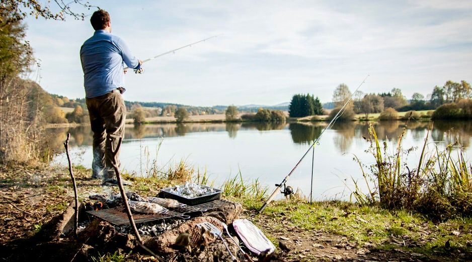 Person fishing at a lake in Czech Republic countryside, with fresh caught zander fillet cooking on BBQ grill in foreground, fishing equipment and tackle box nearby, surrounded by trees and fields under cloudy sky