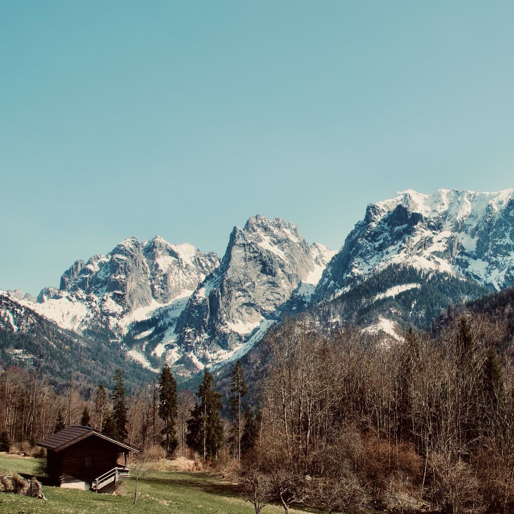 Alpine meadow with traditional wooden hut in Kaisertal valley, Wilder Kaiser mountain range with snow-covered peaks in background, spring landscape in Tyrol Austria April 2018.