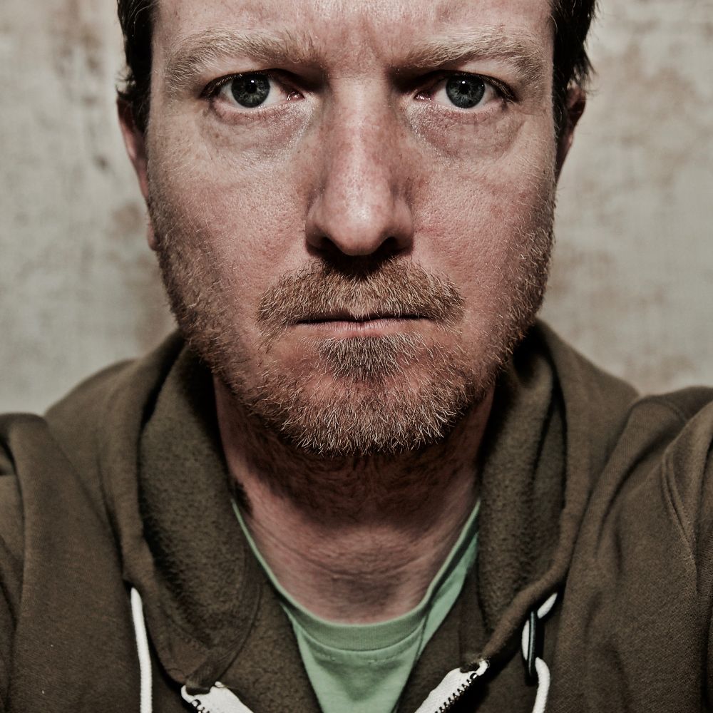 This is a close-up self-portrait photograph showing the face and upper torso of a man with light-colored eyes and short dark hair. He has facial stubble and is wearing an olive green or brown hooded sweatshirt over what appears to be a light green t-shirt. The photo has a neutral, beige-toned background and the subject is looking directly at the camera with a neutral expression. The image has strong detail and is portrait photograph taken in 2017 using a Nikon D70.