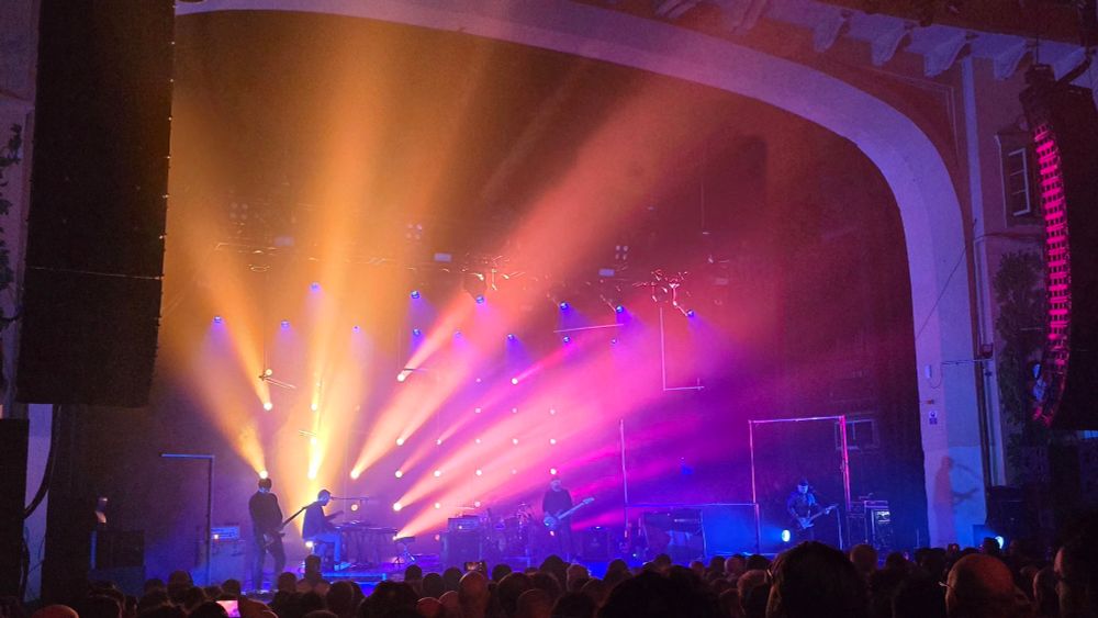 A stage illuminated with bright yellow pink, and blue lights. The post-rock band Mogwai are performing on the stage. 