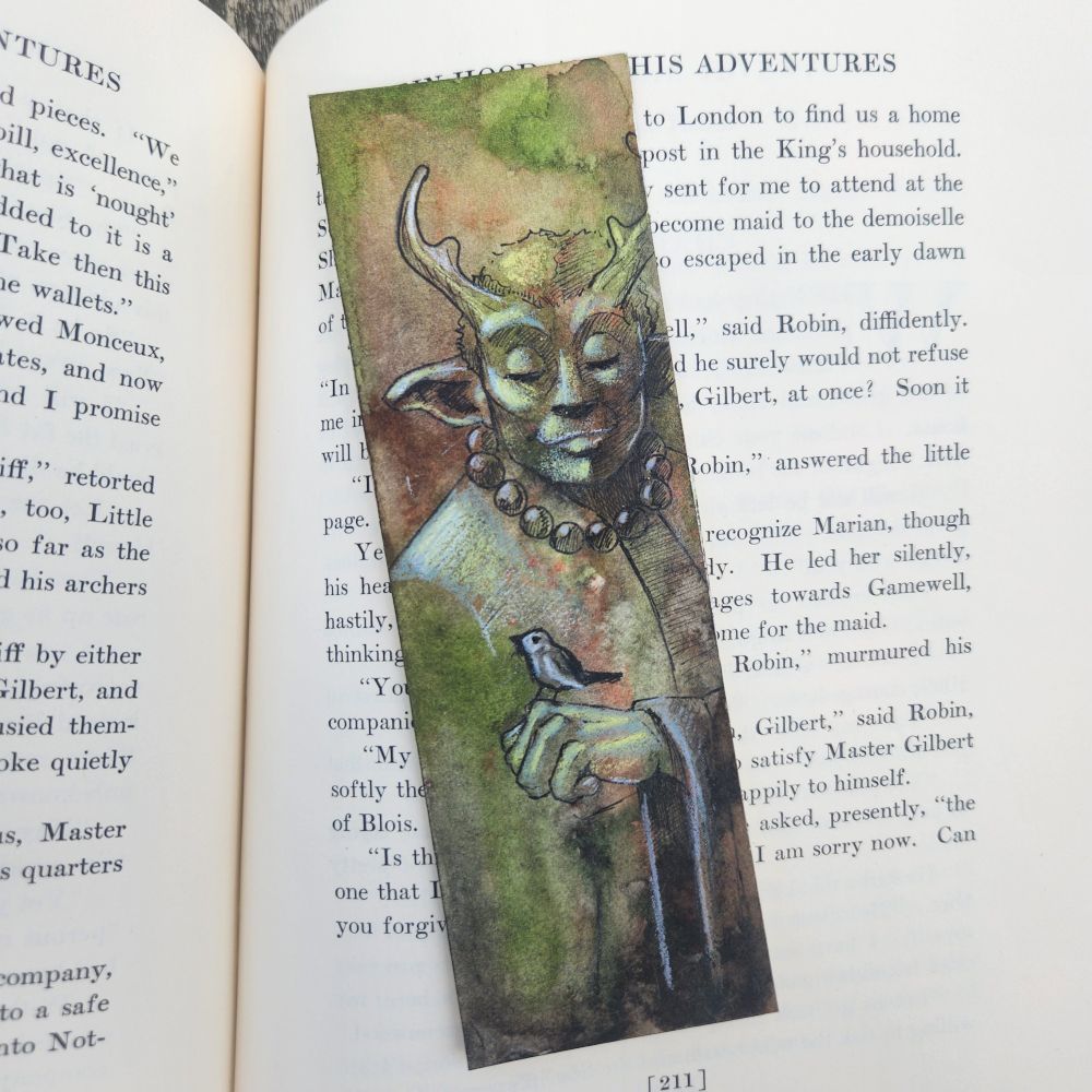 A custom painted bookmark in natural green and brown tones of a serene masculine monkish figure. He has antlers and pointed ears. He considers a small finch like bird perched on his hand