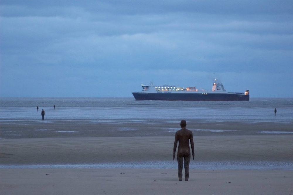 Lots of human figures standing on a beach and in the shallows looking out to sea. There's also a ship in the background 