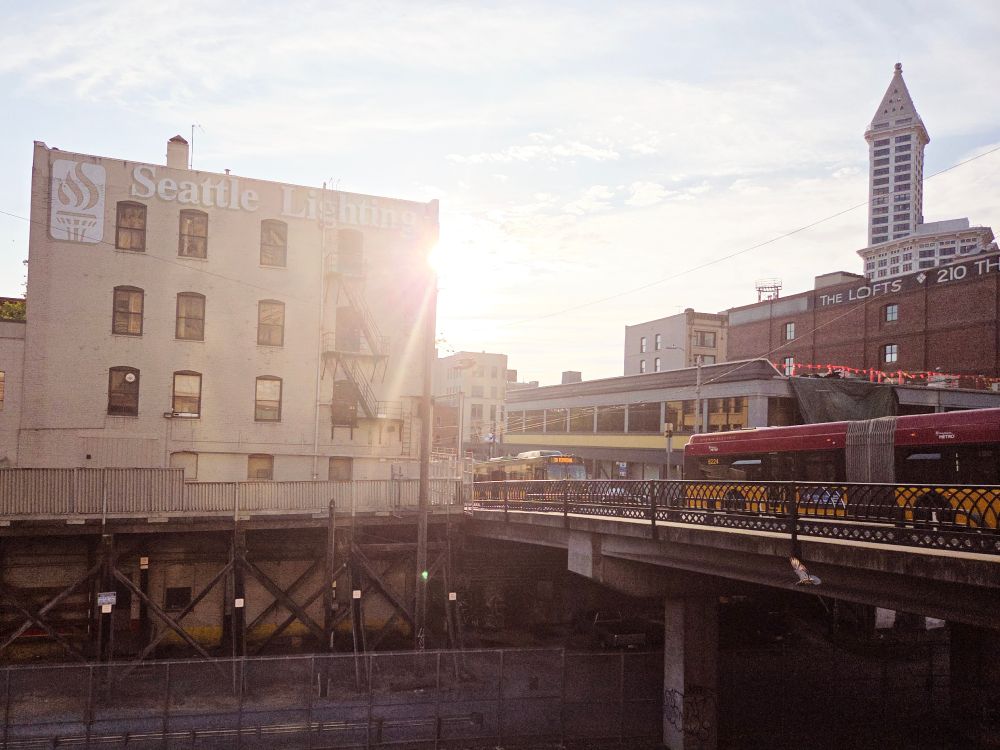 Seattle lighting building with the sun peaking out causing a flare. A bridge over the railroad tracks and the Smith Tower are visible on the right.