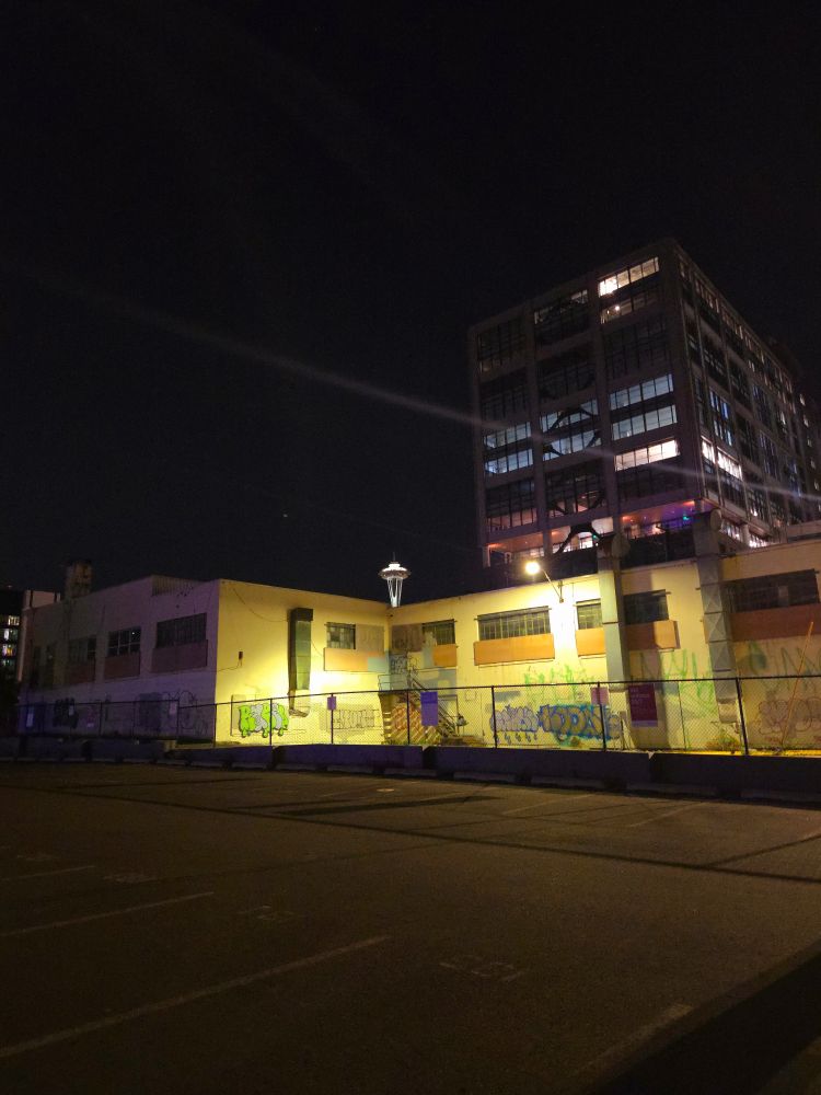 The top of the Space Needle peeking out from behind an unoccupied and graffiti covered building at night. Security lights provide a warm glow, and a high-rise building looms over in the background 