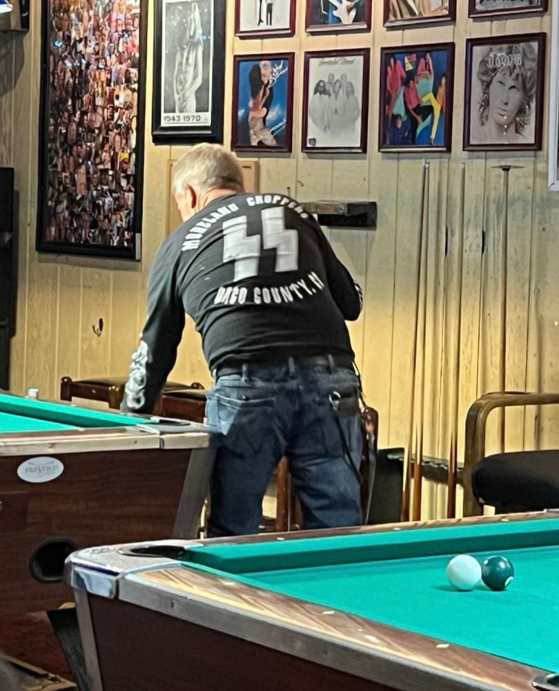 A man stands next to a pool table as he reaches down. The SS logo covers his back with text above and below.