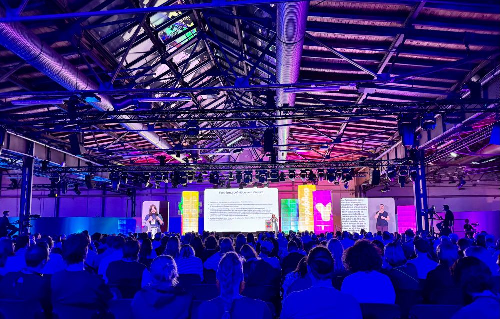 Totale der Bühne mit vielen Zuschauern im Vordergrund von hinten und auf der Bühne Natascha Strobl