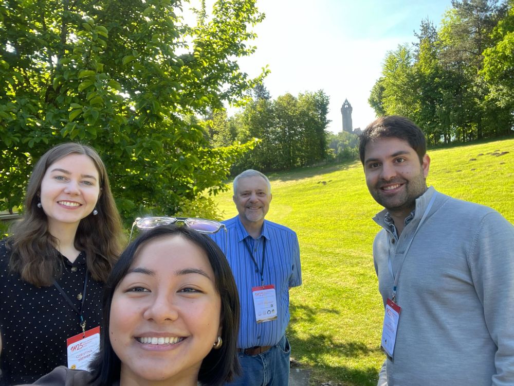 Selfie of four amiling people in front of a leafy background with a medieval tower in the background. 