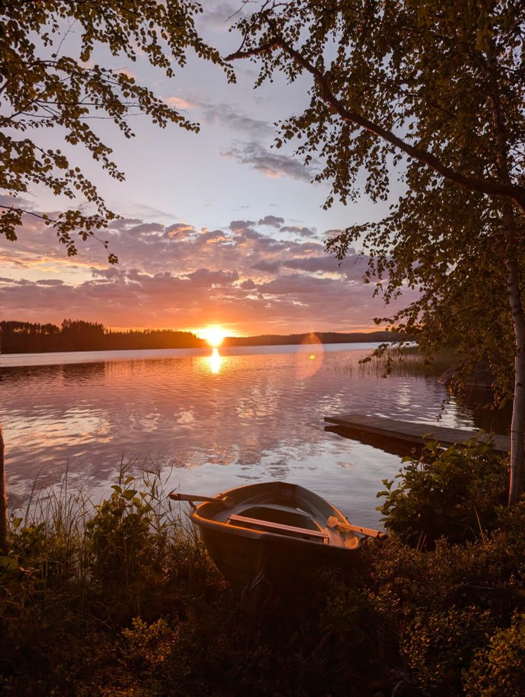Relaxing post sauna evening at the lake near #Sulkava. A rowing boat lying on the lake shore, a pier leading into the lake, and a setting sun on the horizon behind trees.