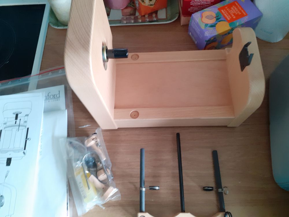 A photo of an unmade up Ashford electric spinning wheel.  The top of the photo sgows the base, the bottom is the flyer. To the left is the instructions and a mini self seal bag of bits. It all sits on a wooden work surface (and a purple tea bag box is visible).