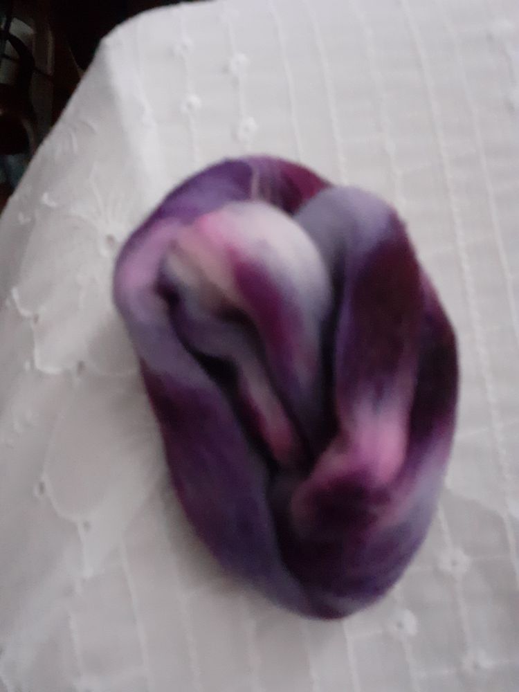 A bit of handdyed fibre. The colours are dark purple, dark lilac and a bit of white. It is sitting on a white fabric background 