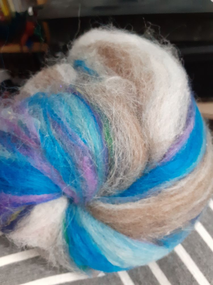 A spiral of fibre with a grey and white striped fabric background.  The batt has a mainly brown and white streak, but there is some blue and purple visible too. 