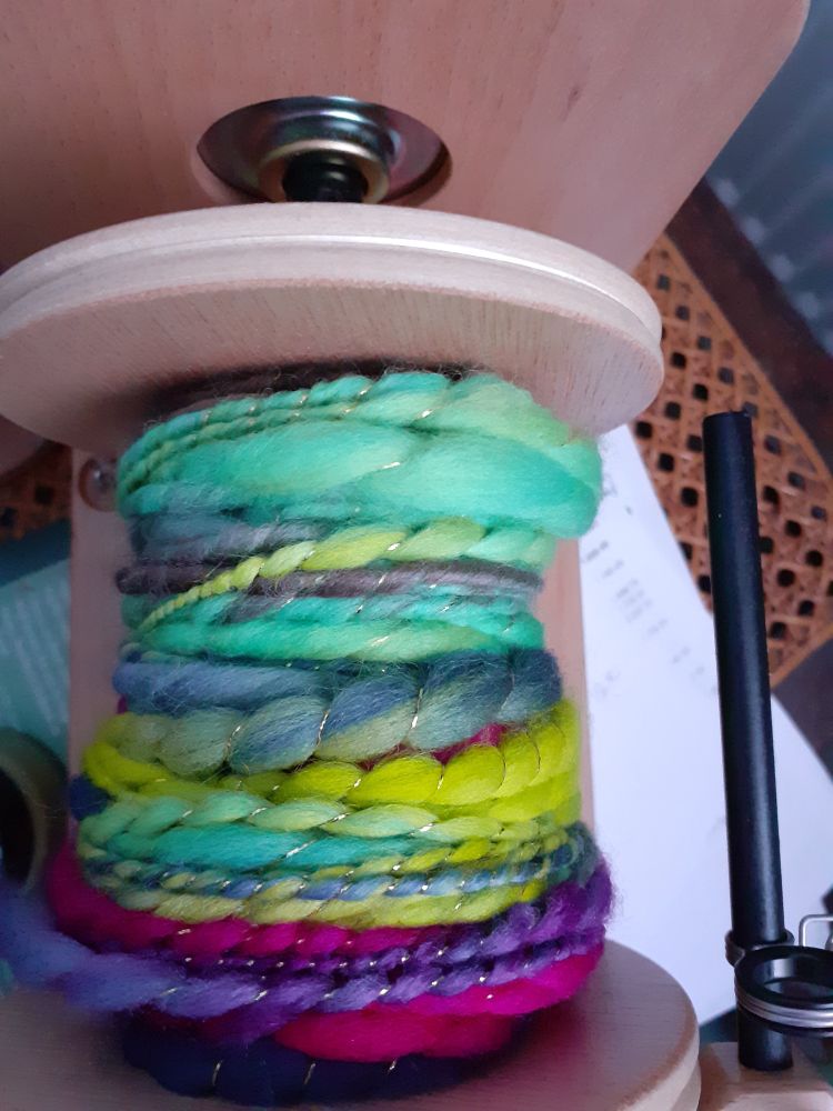 A photo of a wooden electric spinning wheel with a nearly full bobbin of handspun yarn.  The yarn is a thick and thin art yarn with a gold thread plied. The colours are greeny turquoise,  yellow, nay blue and bright pink. The background is a wicker seat with white bit of paper. 