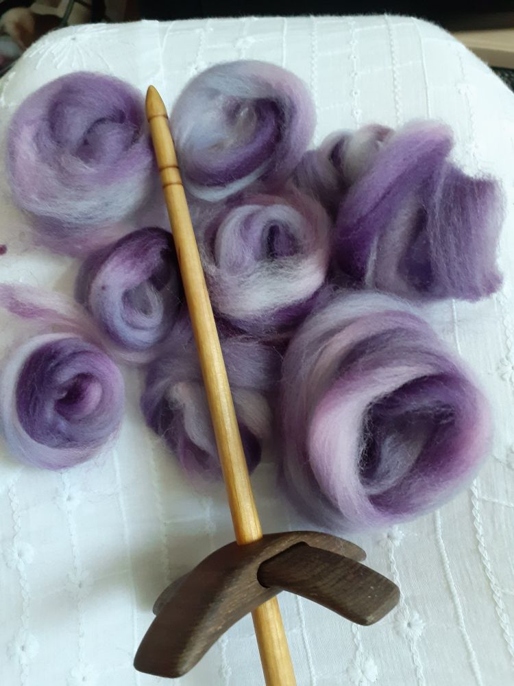 8 spirals of purply fluff sat on a white fabric background with a turkish spindle on top.  The spindle has dark wood arms and a paler pine coloured shaft. 