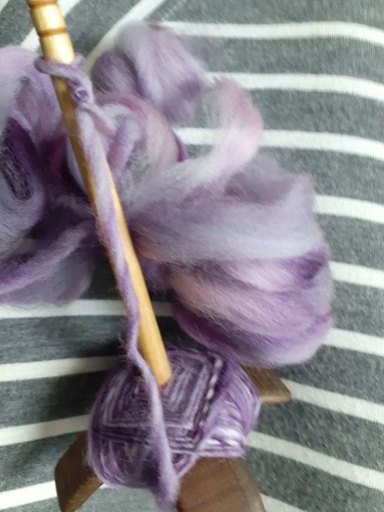 A photo of a wooden turkish spindle with purpley fibre spun and wound on to its arms. The spindle has dark arms and a lighter shaft. There are 3 small bundles of unspun fibre at the top of the photo. Everything is resting on a grey and white striped fabric. 