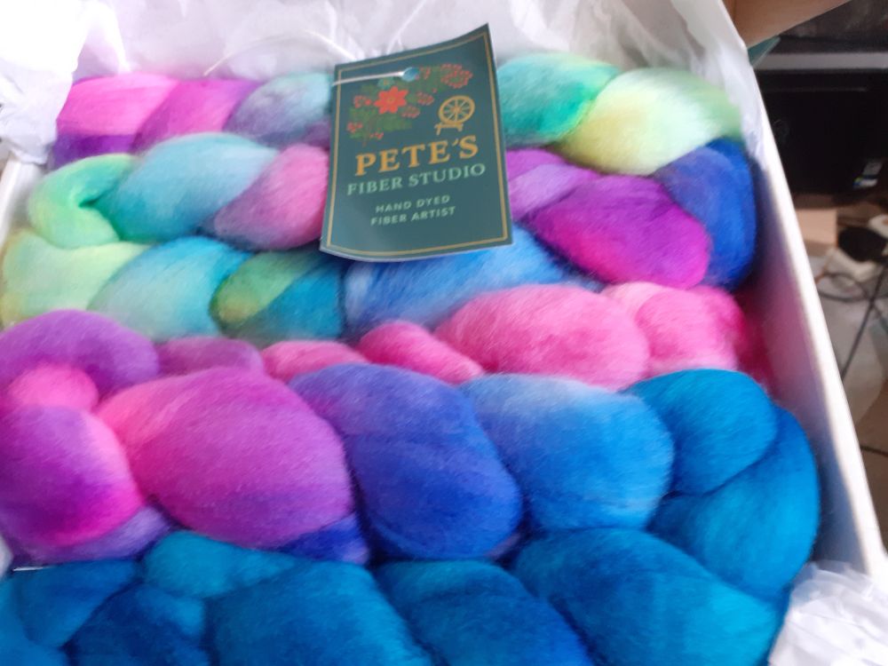 A white box with kid open revealing 2 handdyed braids of fibre. This fibre is Polwarth (sheep). Both braids are plaited and folded in half. The top braid is a pastel rainbow with yellow, green, blue and pink.  The bottom braid is pink into magenta into blue. There is a dark green business card on top of the top braid.