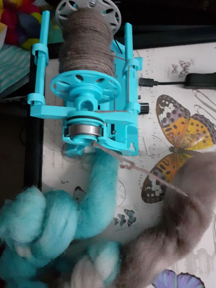 A photo of a half spun small braid of fibre. I am spinning this fibre on my turquoise 3d printed eew nano e-spinner. This sits on a lap tray with a butterfly motif. The fibre is half pale brown and half turquoise. 