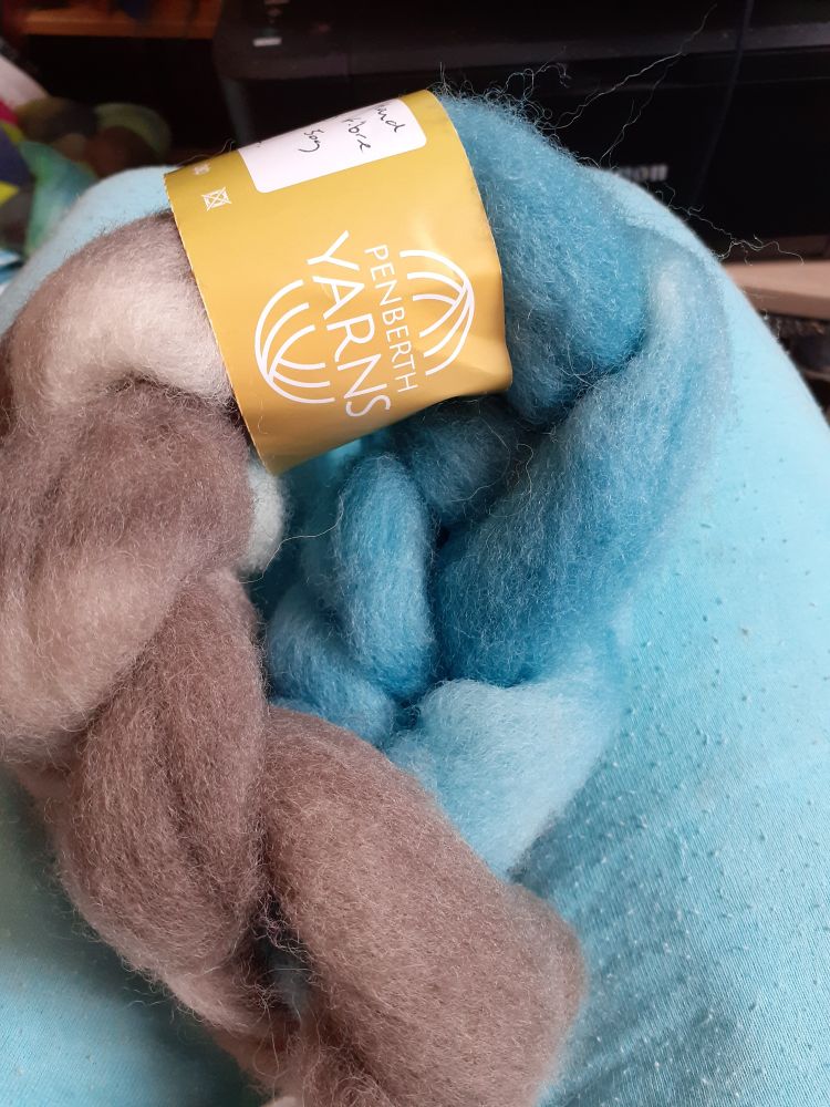 A plaited small braid of fibre in a spiral. Sitting on a pale turquoise fabric background.  The fibre has a yellowy/ orange ball band with the dyer's brand name - Penberth Yarns (a Cornish seller). The fibre is in 2 colours brown and turquoise. 