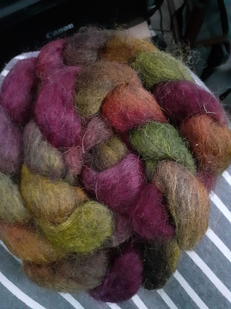 An autumnal handdyed braid of fluff plaited and in a spiral.
The braid sits on a grey and white striped fabric  