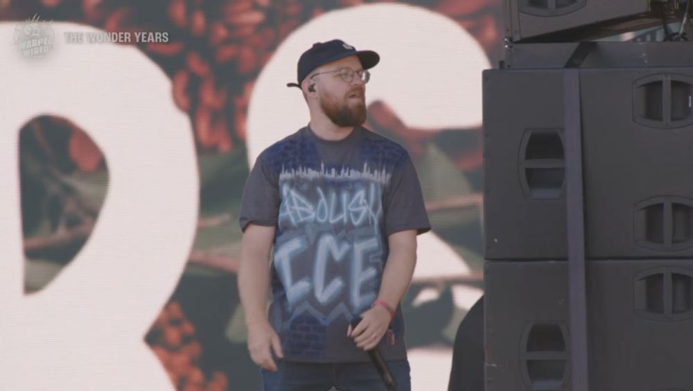 White bearded man with glasses, Dan Campbell of The Wonder Years, on stage next to some speakers. He is wearing a shirt with an airbrushed design that says "ABOLISH ICE" on it.
