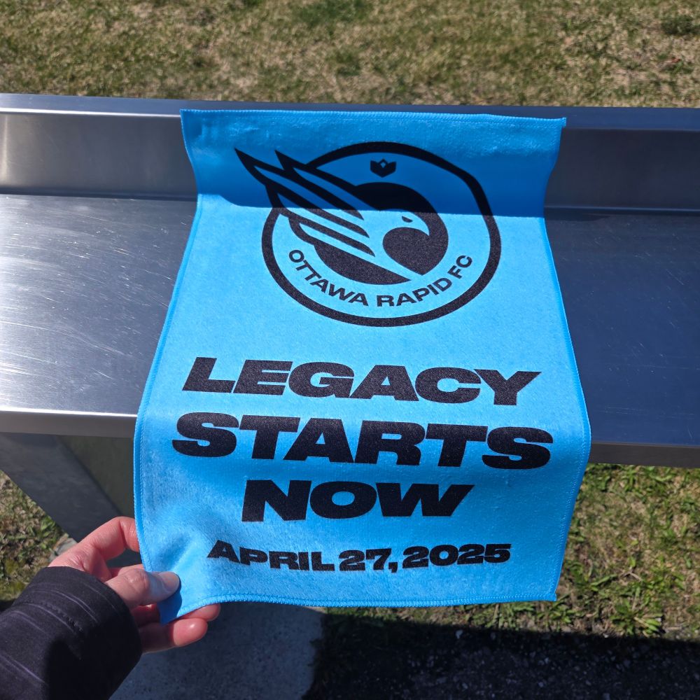 An Ottawa Rapid FC towel, light blue with the logo of a bird in half a heart, the text in black reads "LEGACY STARTS NOW" above today's date in black April 27, 2025.