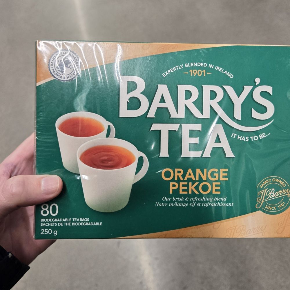 My hand is holding a box of Barty's Tea, a gold and green package announcing "Barry's Tea" with "Orange Pekoe" written below it beside two whutw mug of steaming Tea 