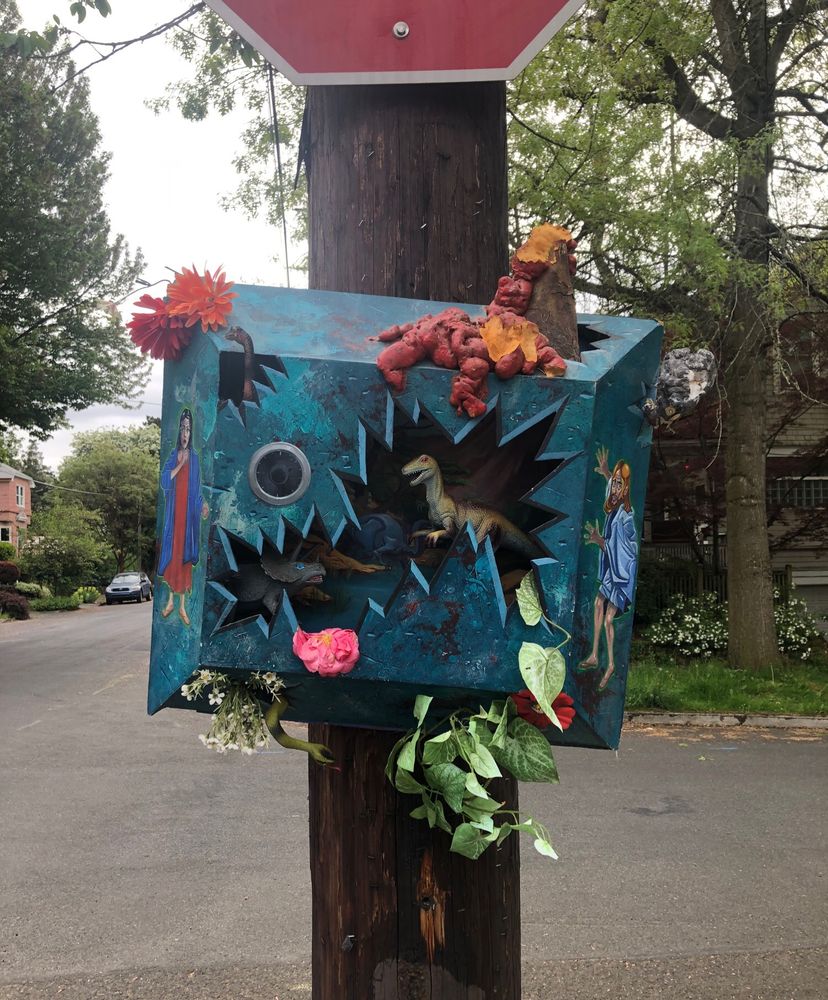 a diorama attached to a power pole on the street with dinosaurs and painted Jesus/catholic saint looking characters on the sides