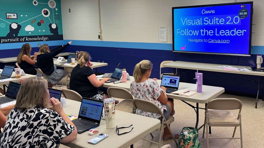Teachers seated at laptops in a classroom during a Canva training session. Presentation screen shows "Visual Suite 2.0 – Follow the Leader." Educators actively participating in design-based professional development.