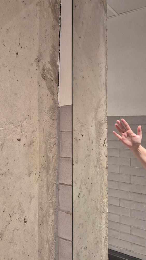 Foto a un ángulo que se forma entre una pared de hormigón y un espejo grande. Se ven reflejadas las baldosas de la pared trasera, grises. Hacia la mitad asomada una mano con la palma extendida hacia delante. No aparece el cuerpo al que va conectada (que es el mío).