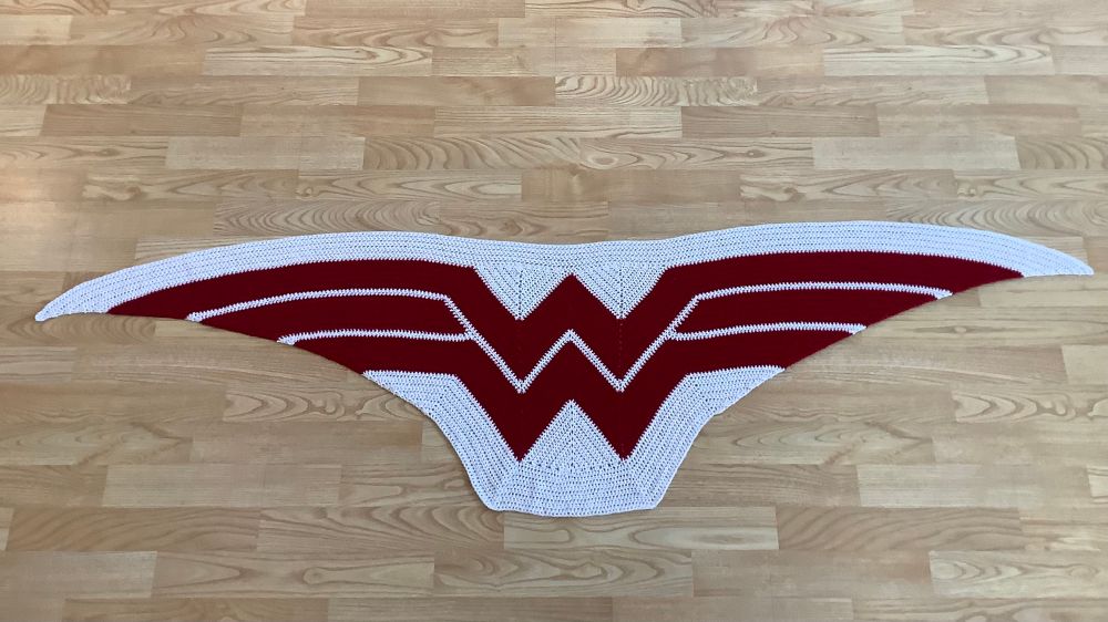 Pictured here is a finished crochet shawl lying on a wood floor. The shawl is red and white, with Wonder Woman’s double W symbol worked into the design. 