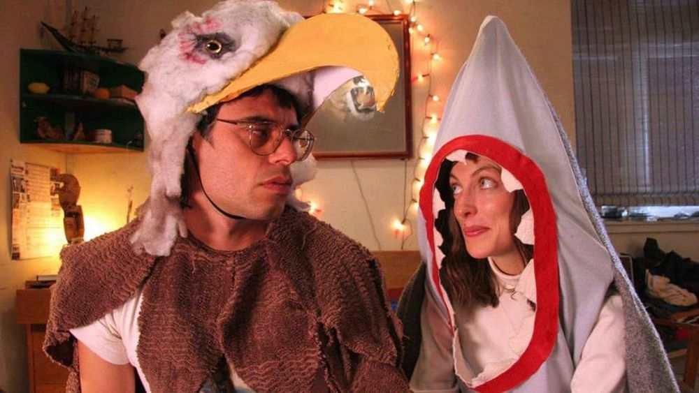 Still from the 2007 comedy movie EAGLE VS SHARK. A woman in a shark costume and a man in an eagle costume are sitting on a sofa at a house party.
