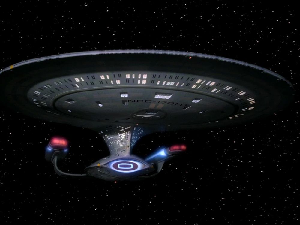 Die USS Enterprise aus Star Trek The Next Generation von schräg vorne. Ein rundliches Schiff. Ein kleiner, ovaler Rumpf, links und rechts zwei nach oben gebogene Triebwerkspylonen. Ein schmaler Hals, der nach oben weiter wird und in einer runden Untertassensektion übergeht. Im Hintergrund ein Sternenhimmel.