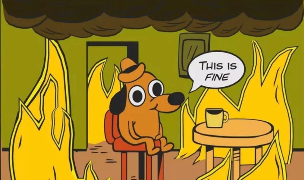 Orange cartoon dog with a hat saying, “This is fine,” while sitting on a red chair surrounded by flames. 