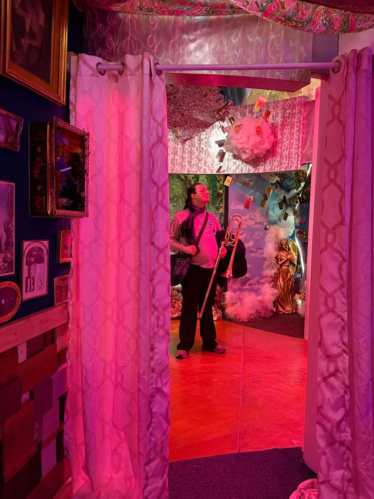 A person stands with a trombone in an artistic display. They are framed by white curtains and the lighting is a soft pink. 