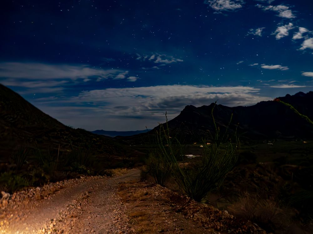 A peaceful nighttime landscape of the Arizona desert under a clear, star-filled sky. Silhouetted mountains frame the scene, and wispy clouds catch faint moonlight. A narrow dirt road, softly illuminated by vehicle headlights, curves gently through sparse vegetation, conveying tranquility of the desert night