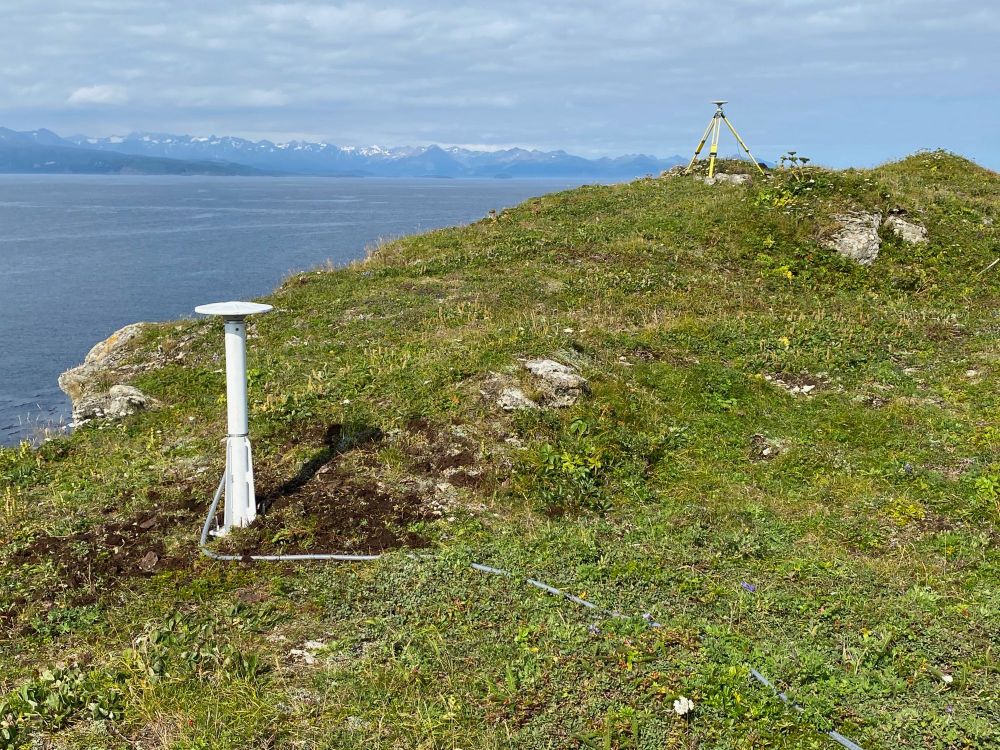 A while cylindrical antenna mast with a white GNSS antenna on top sits on a grassy ridge.  In the distance, a yellow tripod holds another GNSS antenna.  Ocean and mountains stretch off in the distance.  This campaign GNSS site (yellow trip) and continuous GNSS site (white mast) were installed after the 2021 M8.2 Chignik earthquake offshore the Alaska Peninsula.