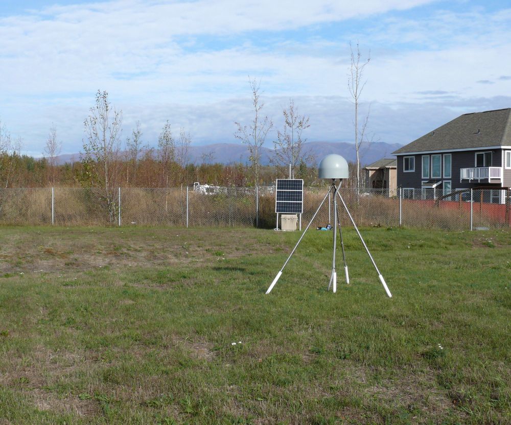 A GNSS antenna mast consisting of three legs stretched out and one center pole and with a gray antenna radome sitting on top is installed on a green grassy area.  Behind the mast is an array of three solar panels with a large tan plastic case that contains batteries, a GNSS receiver, and other electronics.  A house and a trees fill the background.  This permanent GNSS site was installed after the M7.1 Anchorage earthquake in 2018.
